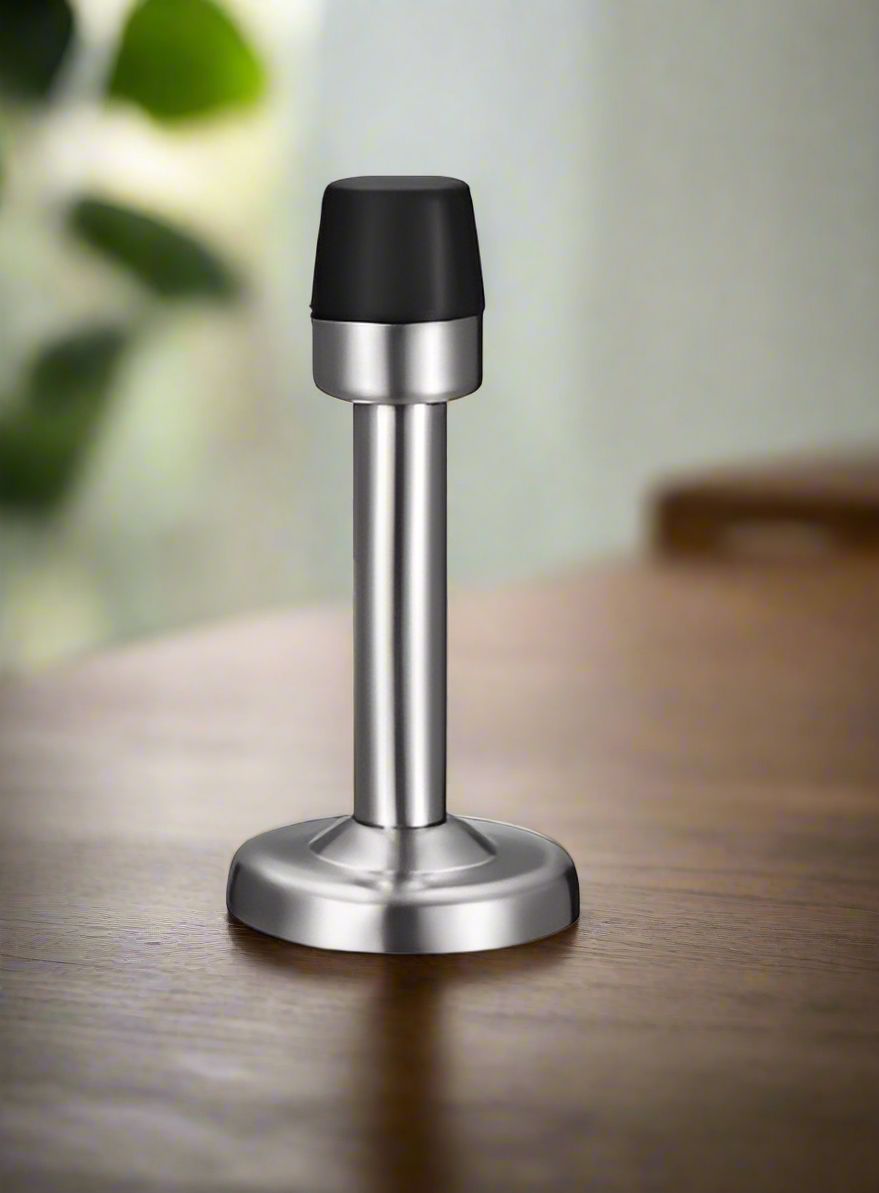 A black and silver wall-mounted door stopper with a rubber tip, installed on a wooden floor.