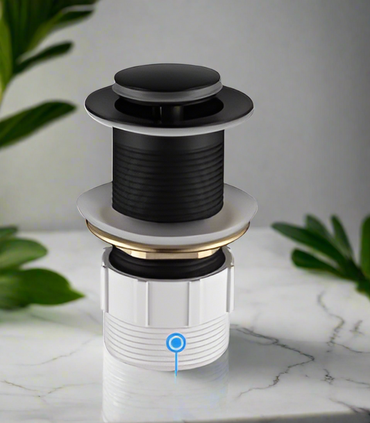 A matt black pop-up basin waste with non-overflow feature, shown against a white marble background with green leafy plants in the backdrop.