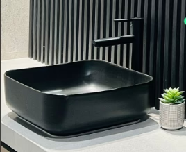 A matte black ceramic above-counter square wash basin displayed in a bathroom setting with a dark vertical striped wall in the background and a small potted plant to the right.