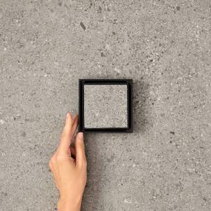 Hand reaching towards a small black square frame on a gray concrete surface